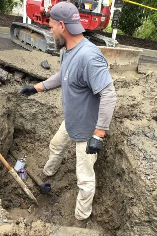 Crew Plumbing plumber in an excavation with a mini excavator behind him